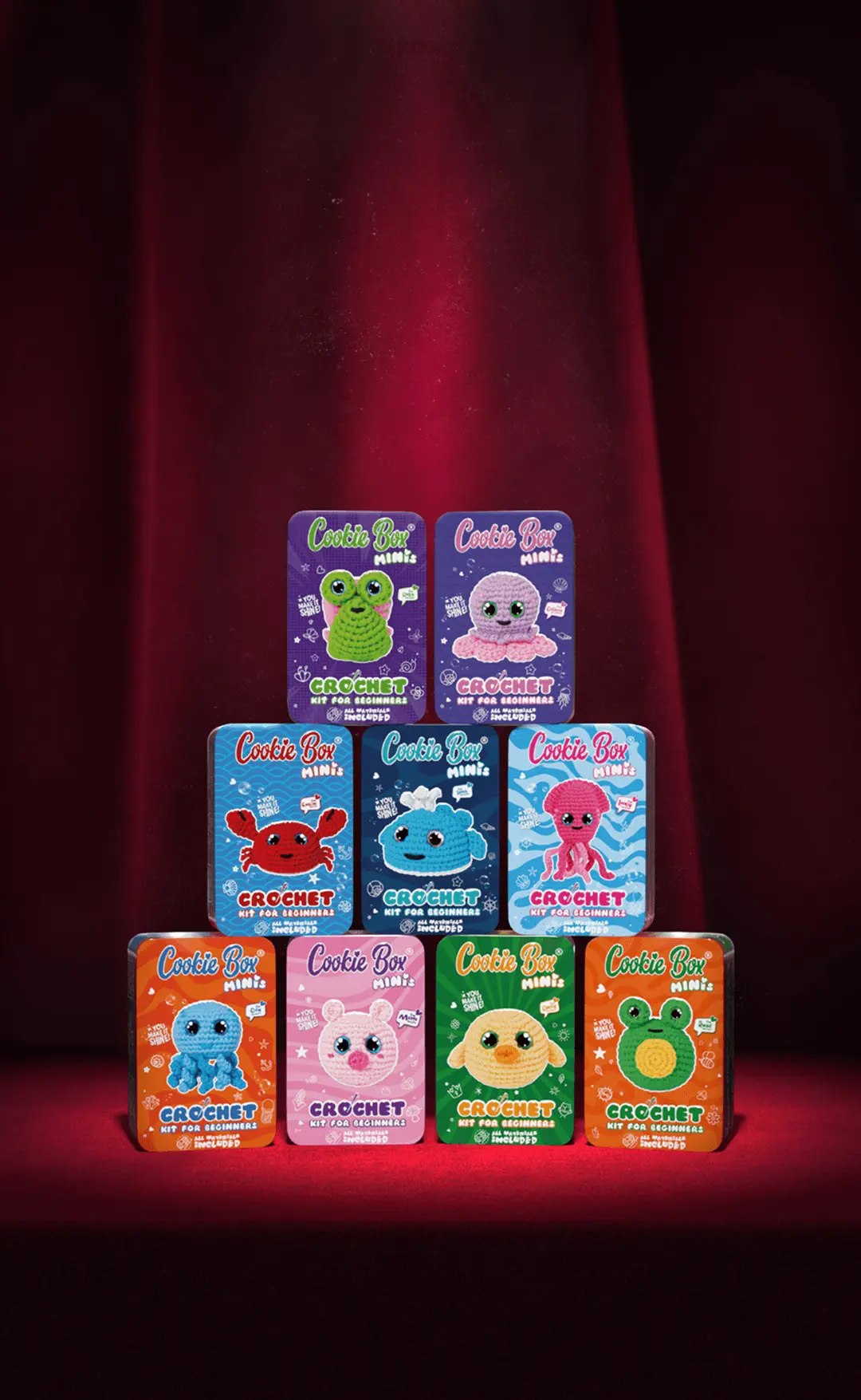 Pyramid of nine Cookie Box Minis crochet kit tins under a spotlight on a red curtain stage.